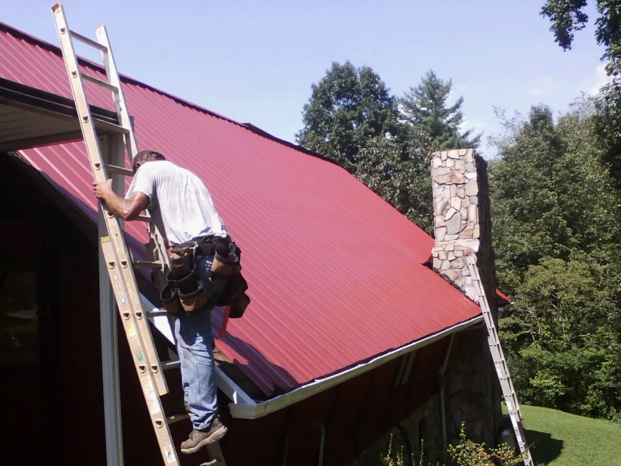 Roofer on a ladder beside a red metal roof and stone chimney — Soffit & Fascia Repair by Roofing Brothers in Bothell